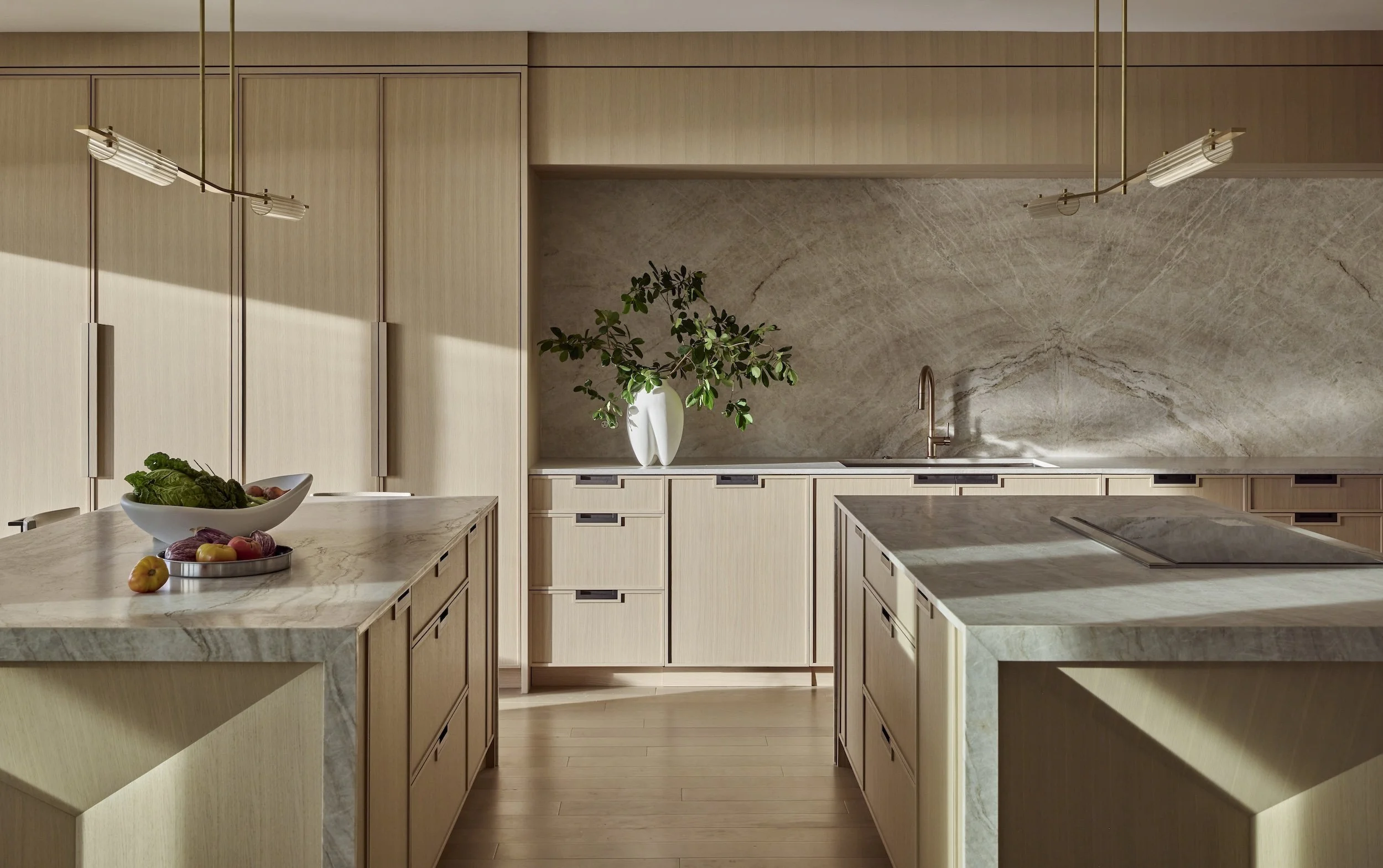 Modern kitchen with wooden cabinets, marble countertops, a large marble backsplash, a sink with a brass faucet, a potted plant in a white vase, and a bowl of vegetables and fruits on the counter.