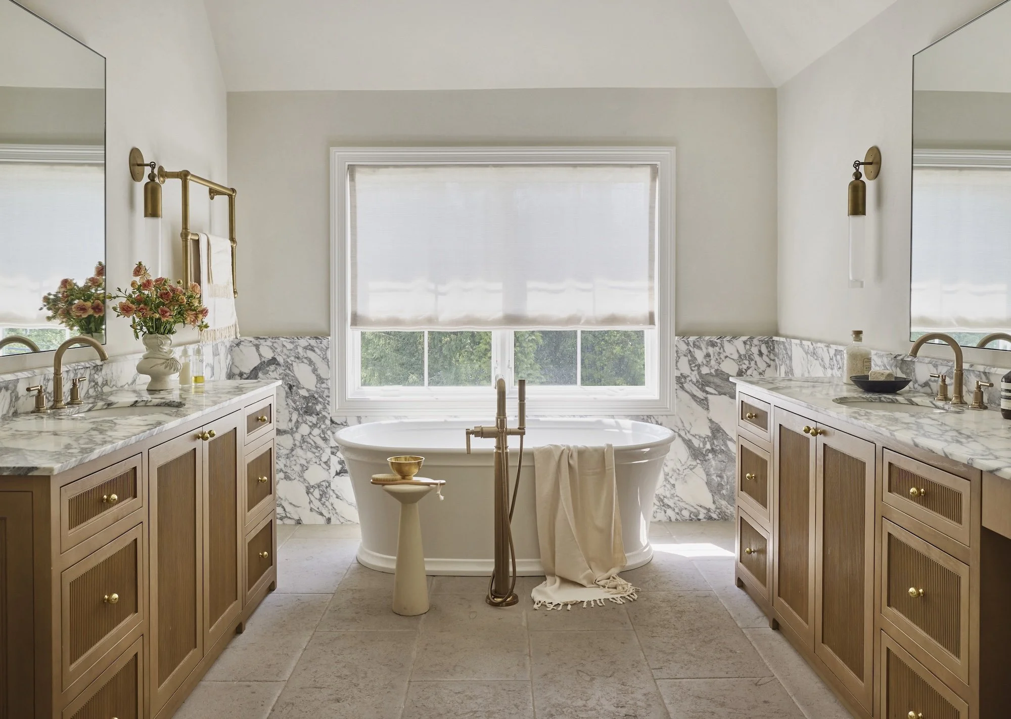 Bright bathroom with a large window, marble countertops, wooden cabinets, a standalone bathtub, and decorative flowers.