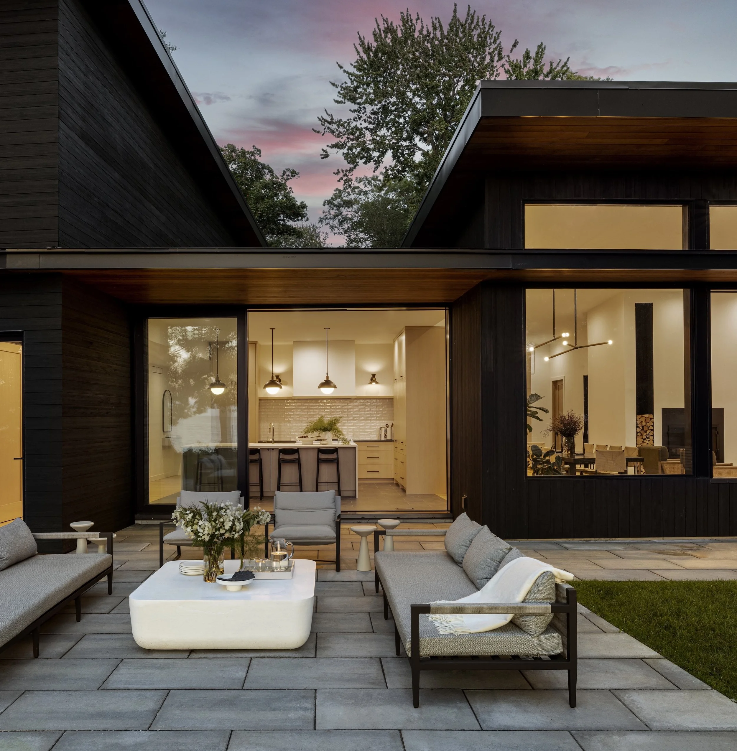 Modern outdoor patio area with light gray cushioned sofas, a white coffee table with decorative items, and a black armchair, all on gray wooden decking. There are small side tables, a bouquet of white flowers on the coffee table, and a lush green lawn in the background. The house features large glass windows and a black and wood exterior. The interior of the house is warmly lit, and the sky is a dusk pink with trees visible in the background.