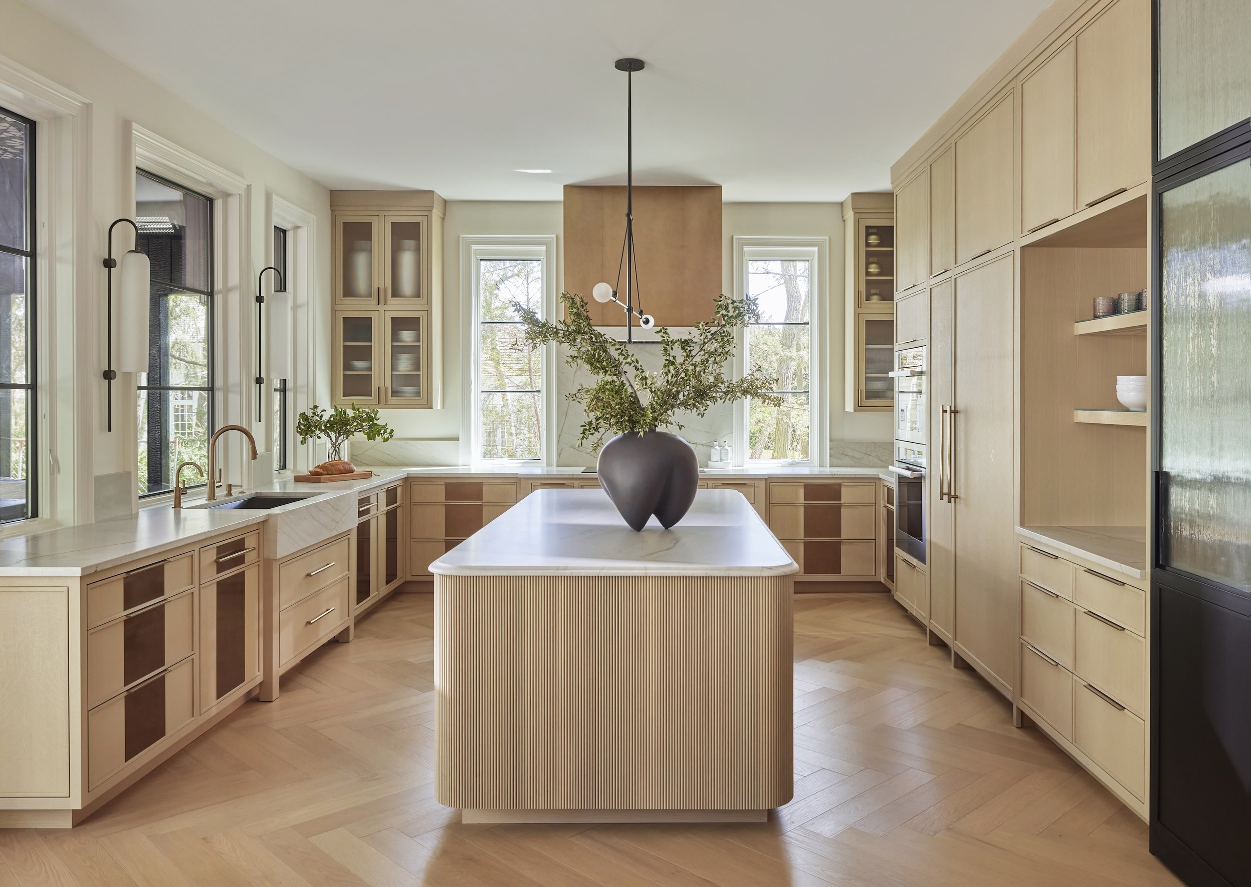 A modern kitchen with light wood cabinetry, large windows, a central island with a vase of greenery, and a minimalist black pendant light hanging from the ceiling.