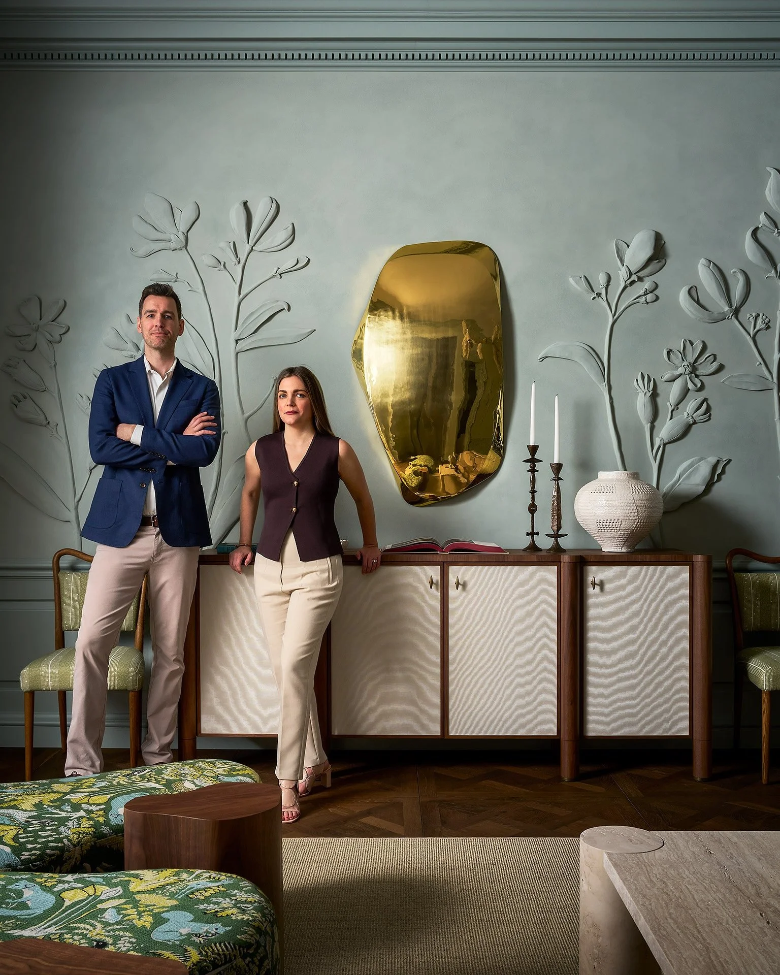 A man and woman standing in a decorated living room with floral wall art, a gold mirror, and modern furniture.