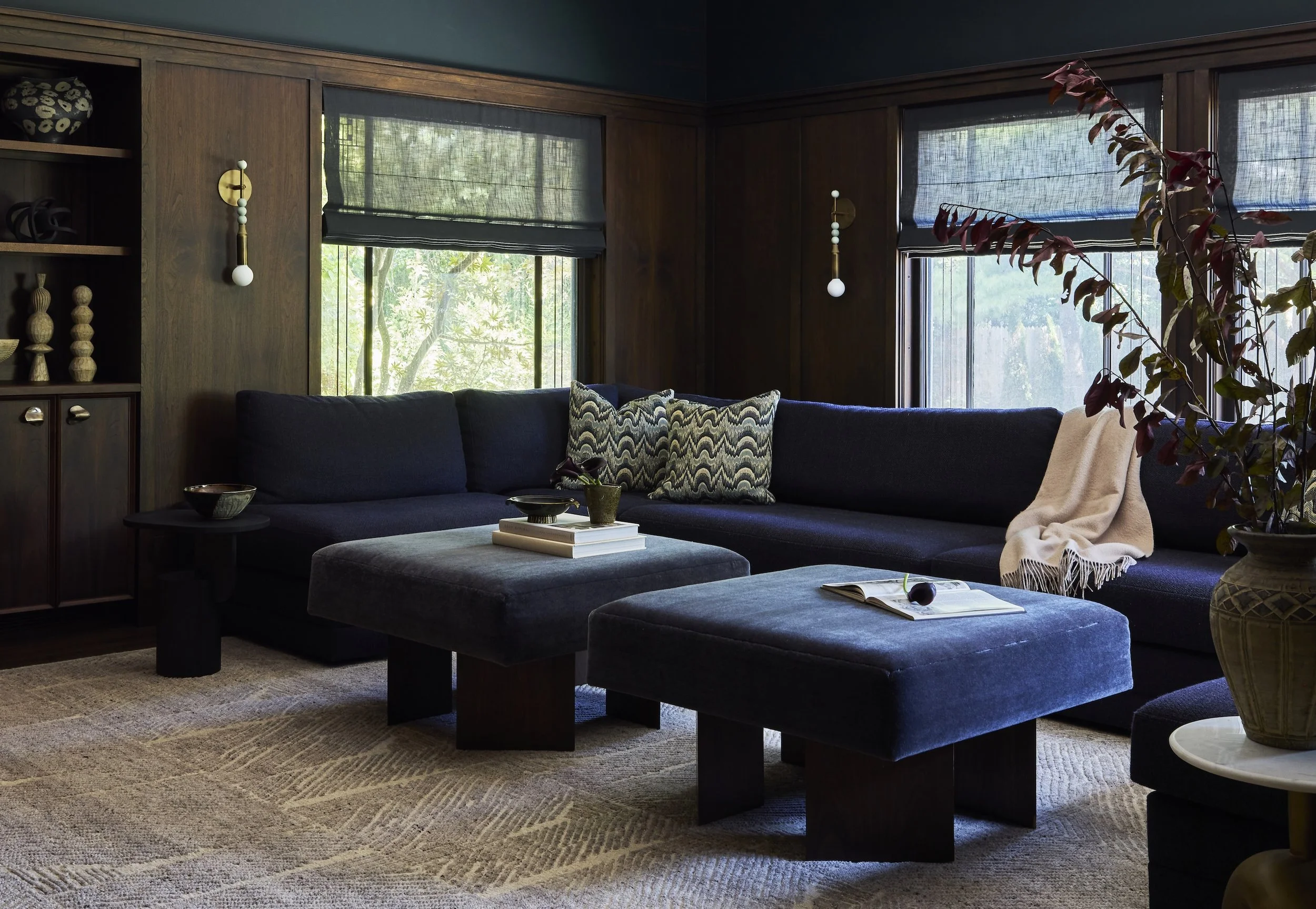 A living room with dark furniture, including a large black sectional sofa with patterned pillows, a dark square ottoman, and a side table with a decorative vase. There are two windows with black Roman shades, wood-paneled walls, and wall-mounted lamps. A large potted plant with reddish leaves is on the right, and a beige throw blanket is draped over the sofa.
