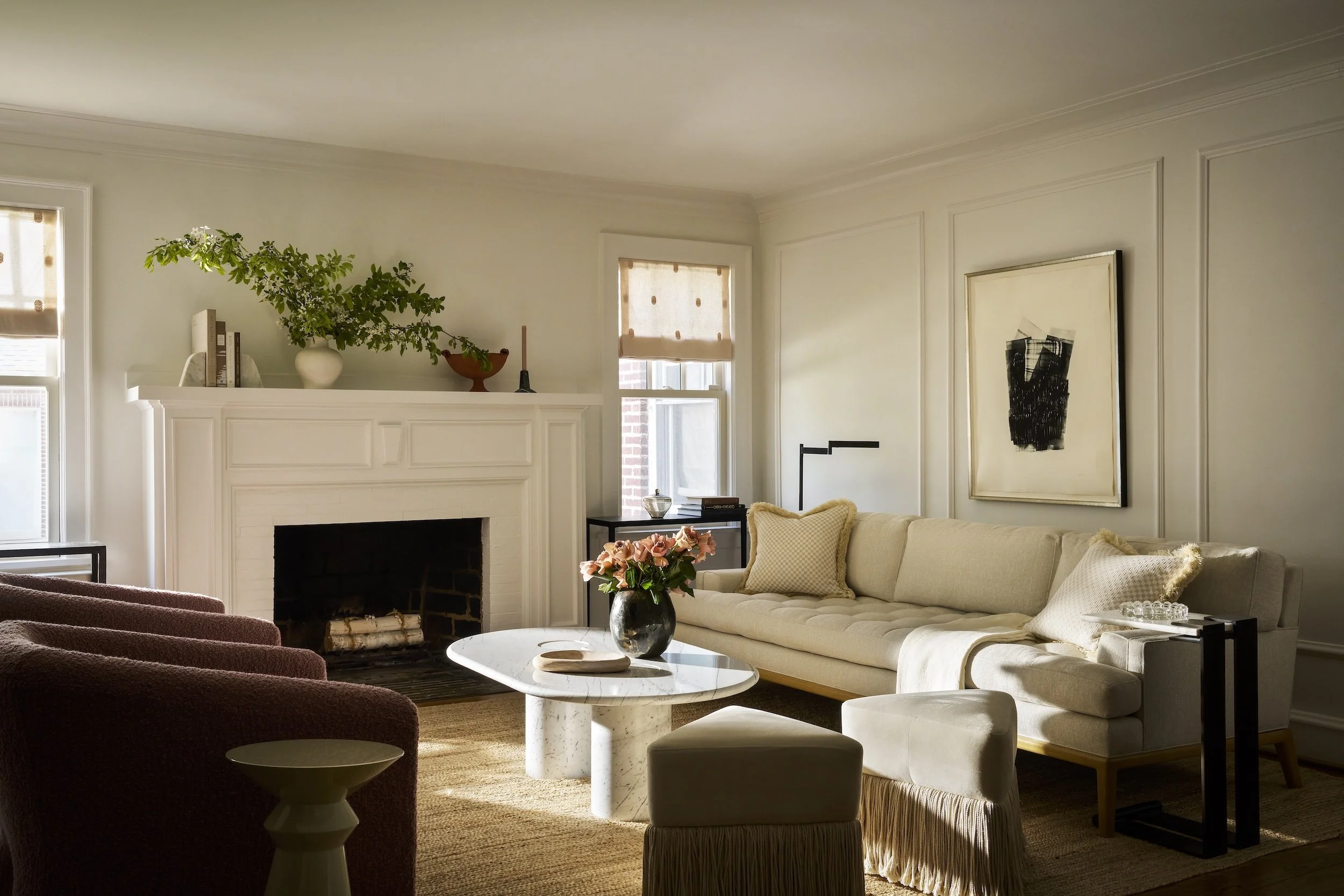 A cozy living room with a white sofa, a wine-colored armchair, and a marble-top coffee table in the center. There is a flower arrangement on the coffee table, and decorative pillows on the sofa. The room features a white fireplace mantel decorated with candles, a vase with greenery, and books. Natural light enters through the windows, which are covered with beige blinds. Artwork hangs on the wall, and the room has a warm, inviting ambiance with neutral tones.