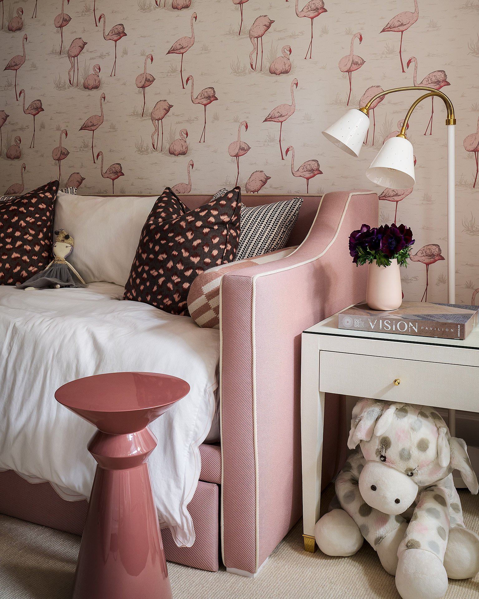 Decorative bedroom with flamingo-themed wallpaper, pink upholstered bed, black and white cushions, a pink side table, a white bedside table with a purple flower arrangement, a white lamp with gold accents, and a large plush giraffe stuffed animal on the floor.