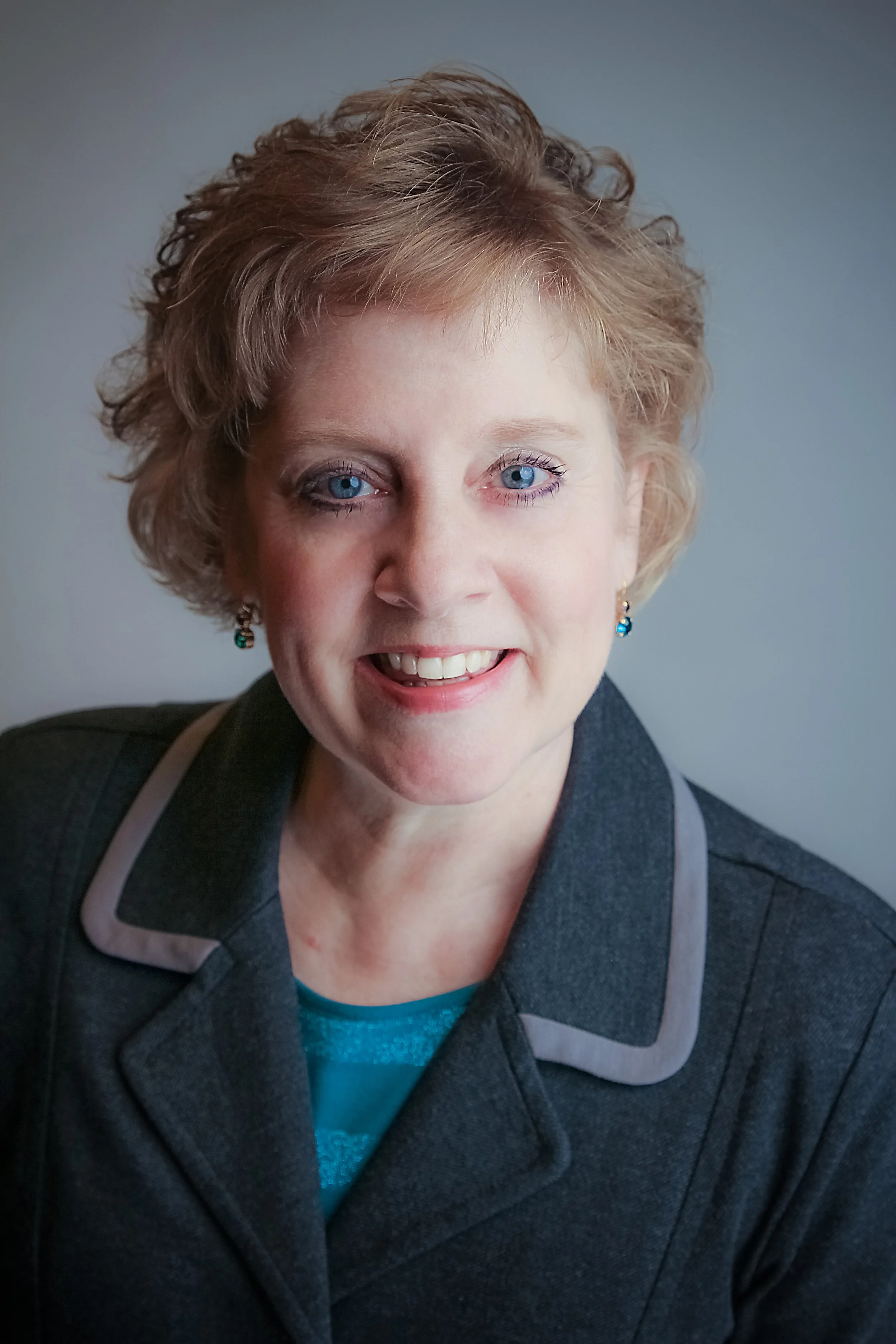 Close-up portrait of a woman with short, curly red hair, blue eyes, and wearing earrings. She is smiling and dressed in a gray blazer and a blue top, posing against a plain background.