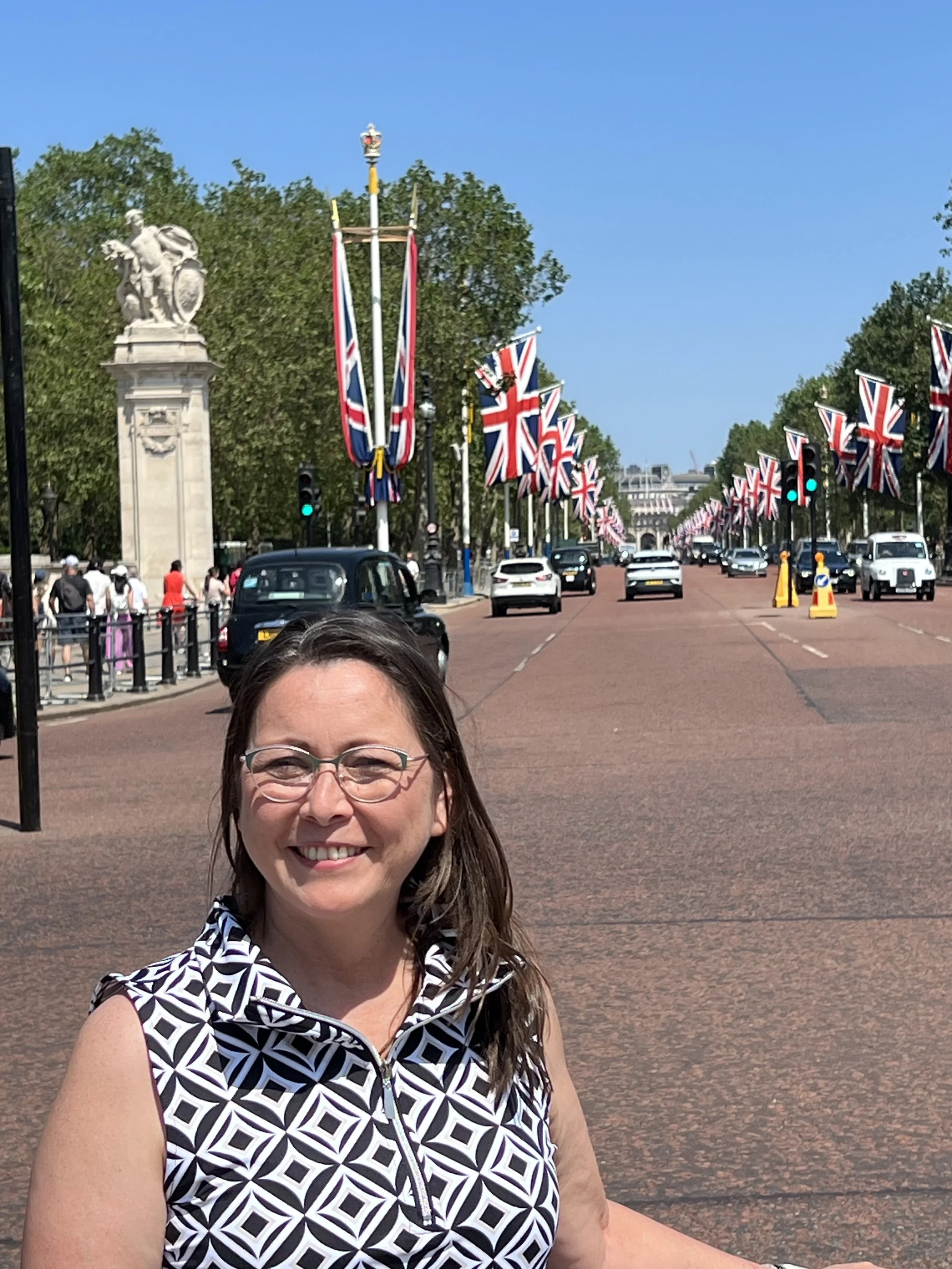 A woman smiling in front of The Mall in London, with Union Jack flags lining the street and a statue on a pedestal visible in the background.