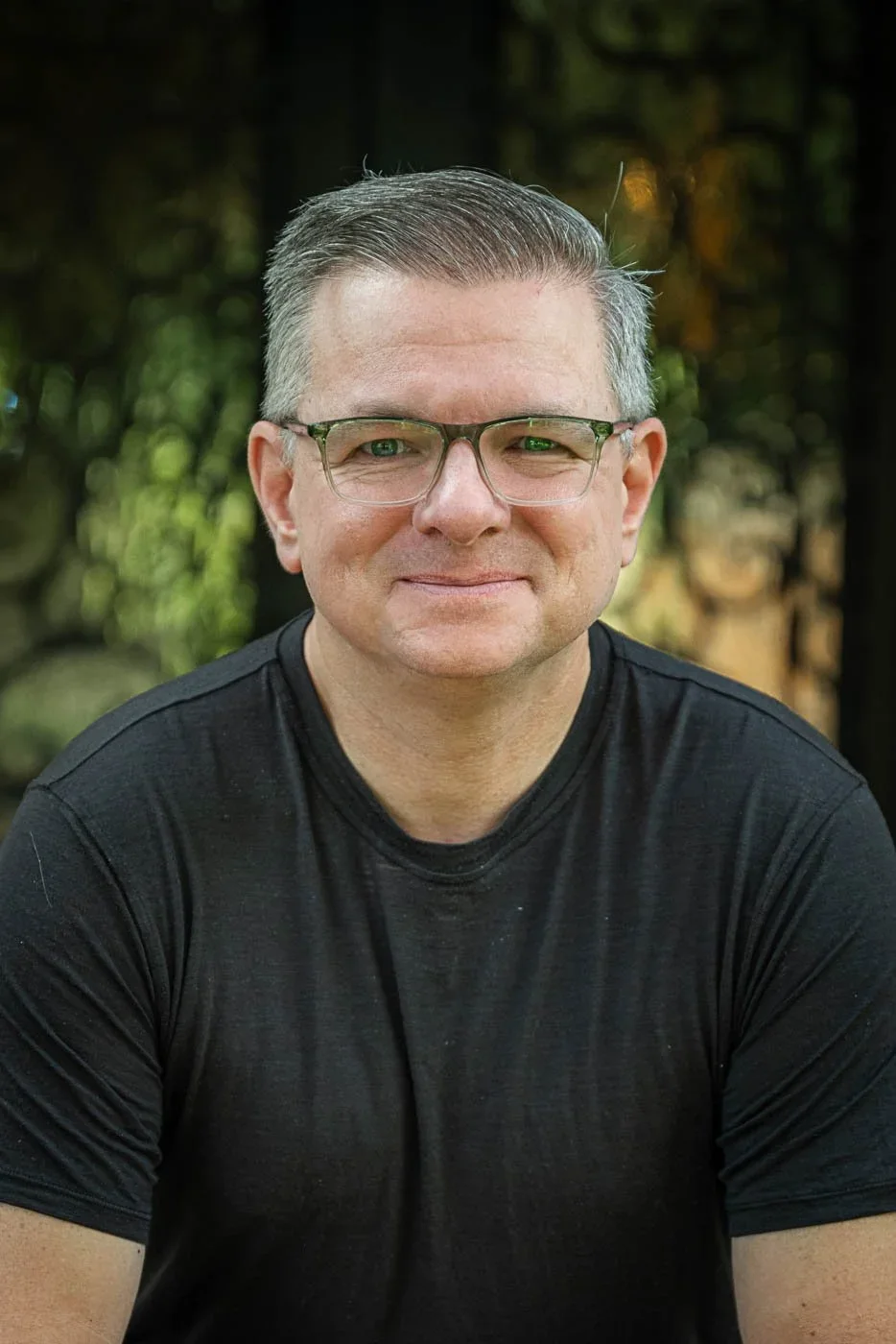 A man with short, gray hair, wearing glasses and a black t-shirt, smiling outdoors with a blurred green and brown background.
