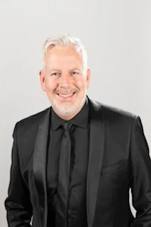 Portrait of a smiling middle-aged man with white hair, wearing a black suit and black shirt, against a light gray background.