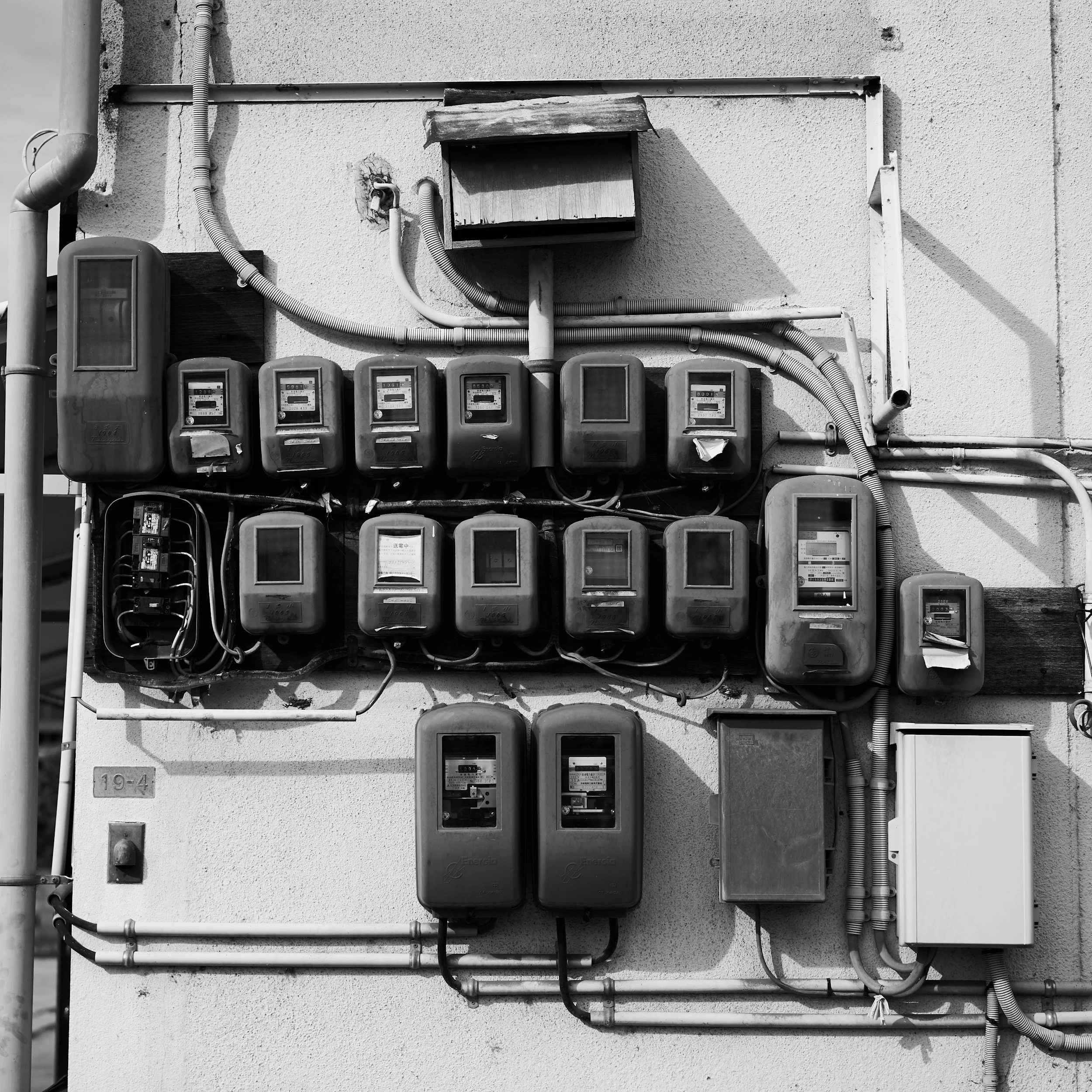 Black and white photo of electrical meters and conduits mounted on an exterior wall, with shadows cast on the wall.