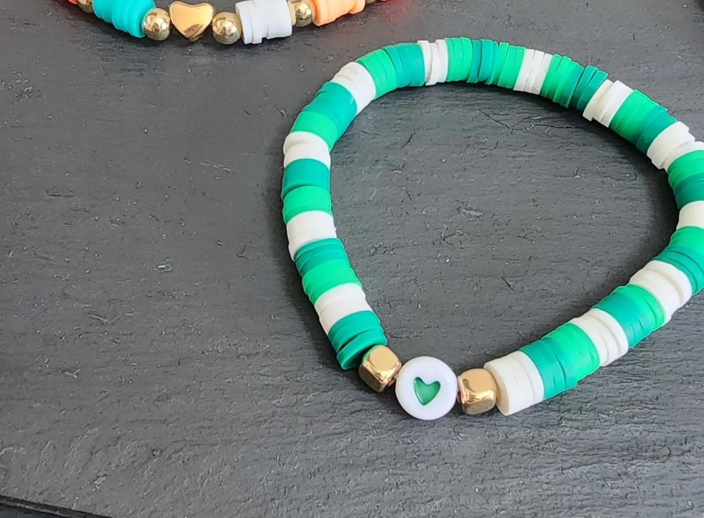 Colorful beaded bracelet composed of blue, white, and gold beads, with a white round bead featuring a green heart symbol.