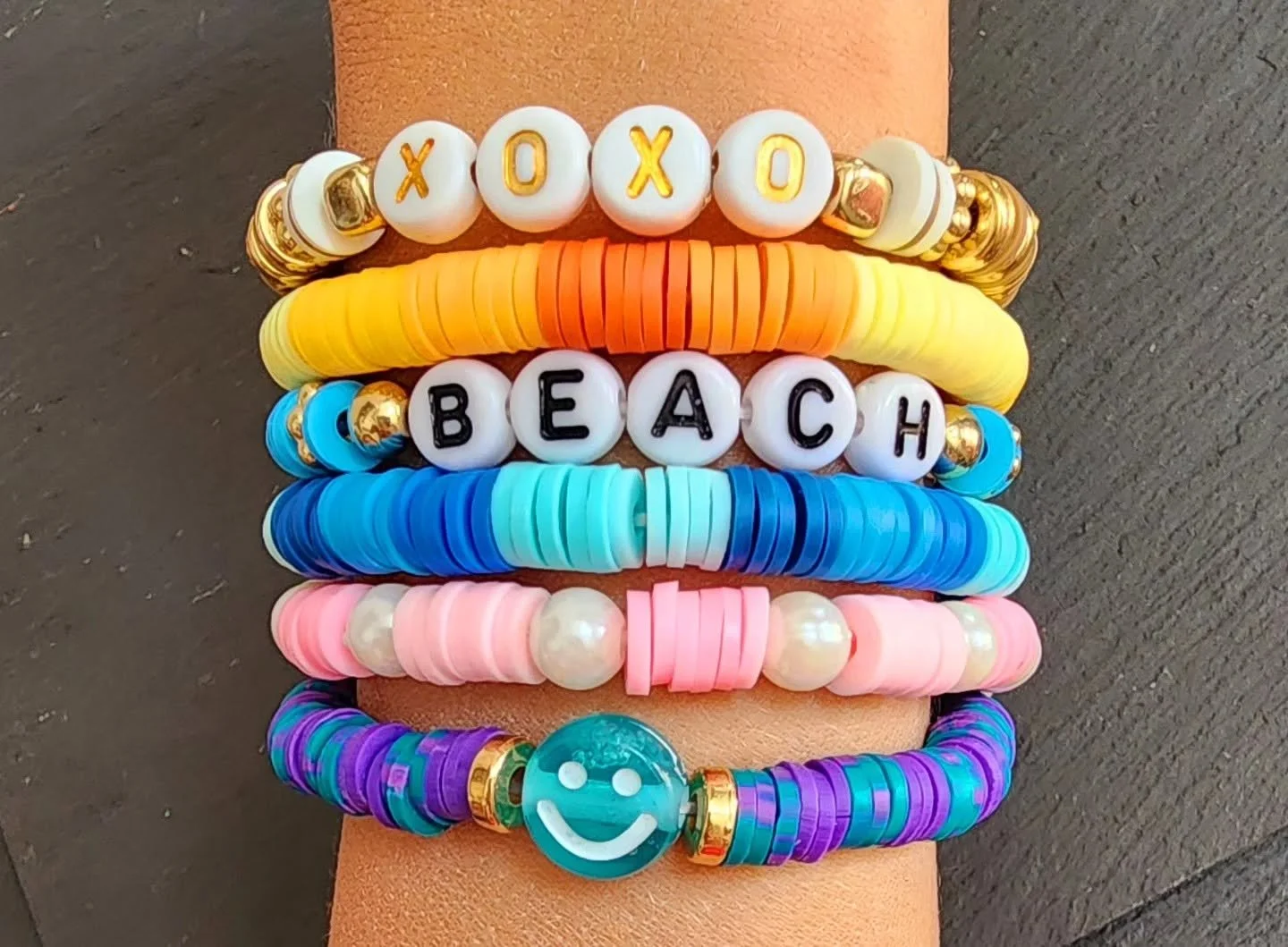 Colorful beaded bracelets arranged on an orange surface spelling out "XOXO BEACH" with a smiley face charm, featuring various beads in white, gold, yellow, orange, blue, pink, and purple.