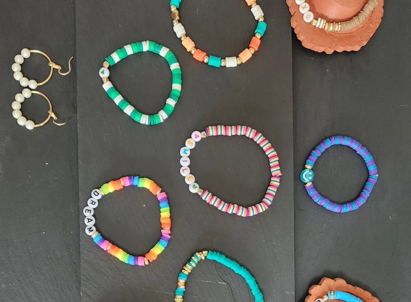 Various colorful bead necklaces and earrings on a black surface. Some necklaces contain alphabet beads spelling words, others are made of multicolored, rainbow, and pastel beads. There are also pearl and gold bead earrings and some stacked clay or terracotta bowls with additional jewelry.