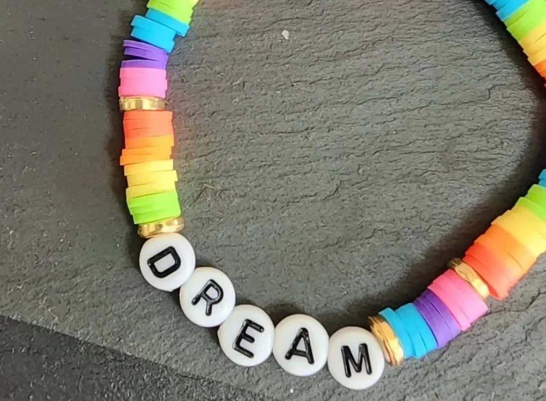 Colorful bracelet made of multicolored beads with white round beads spelling out the word "DREAM" on a gray surface.