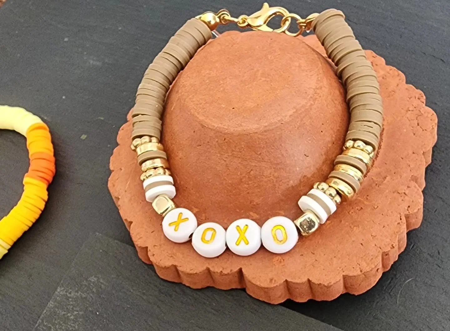 Beaded necklace with white beads spelling out 'XOXO', placed on a round orange terracotta clay block, with another partially visible beaded bracelet nearby on a dark surface.