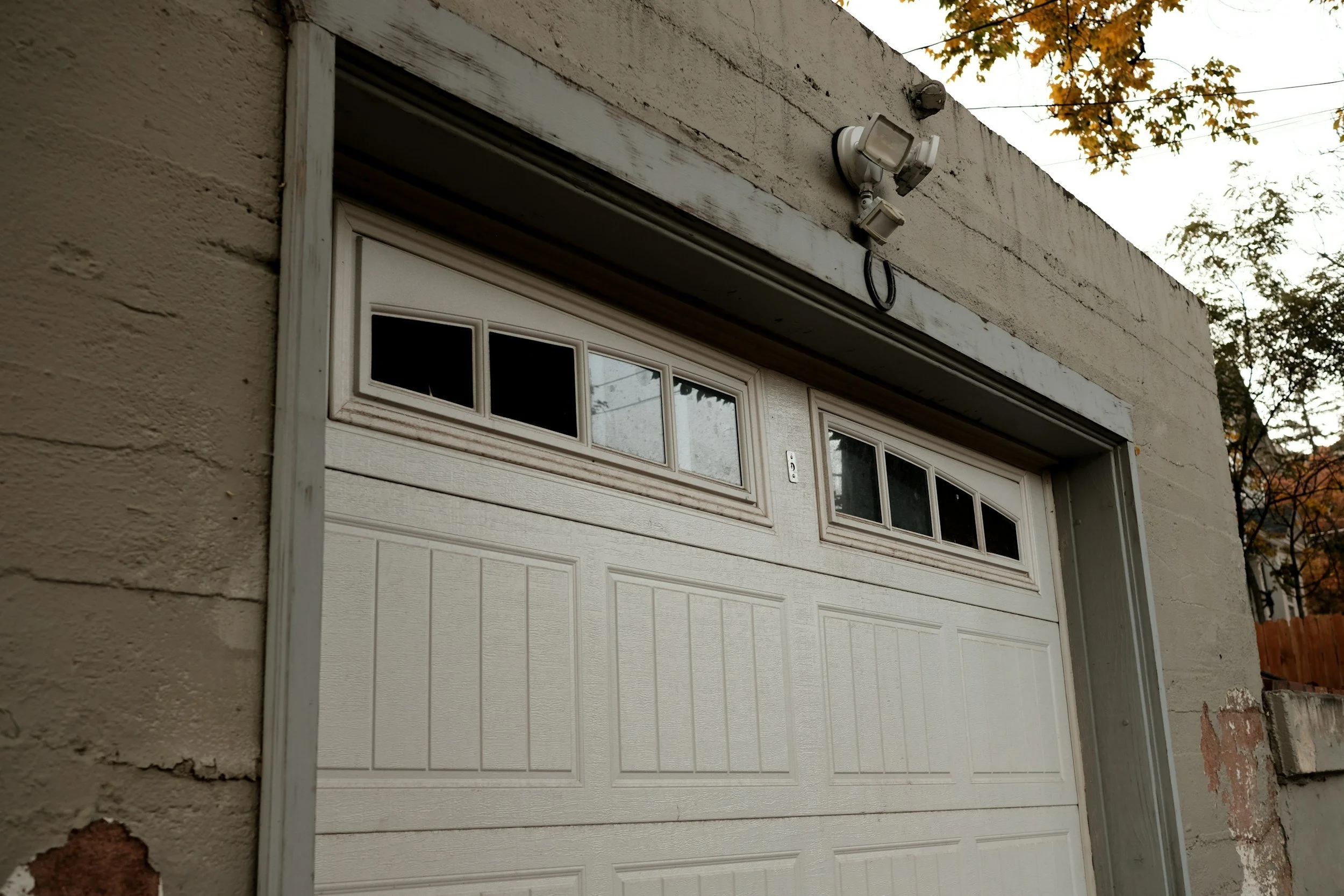 Close-up of a white garage door with small windows at the top, attached to a beige brick building with a weathered wooden trim above, and an outdoor security light mounted on the wall.