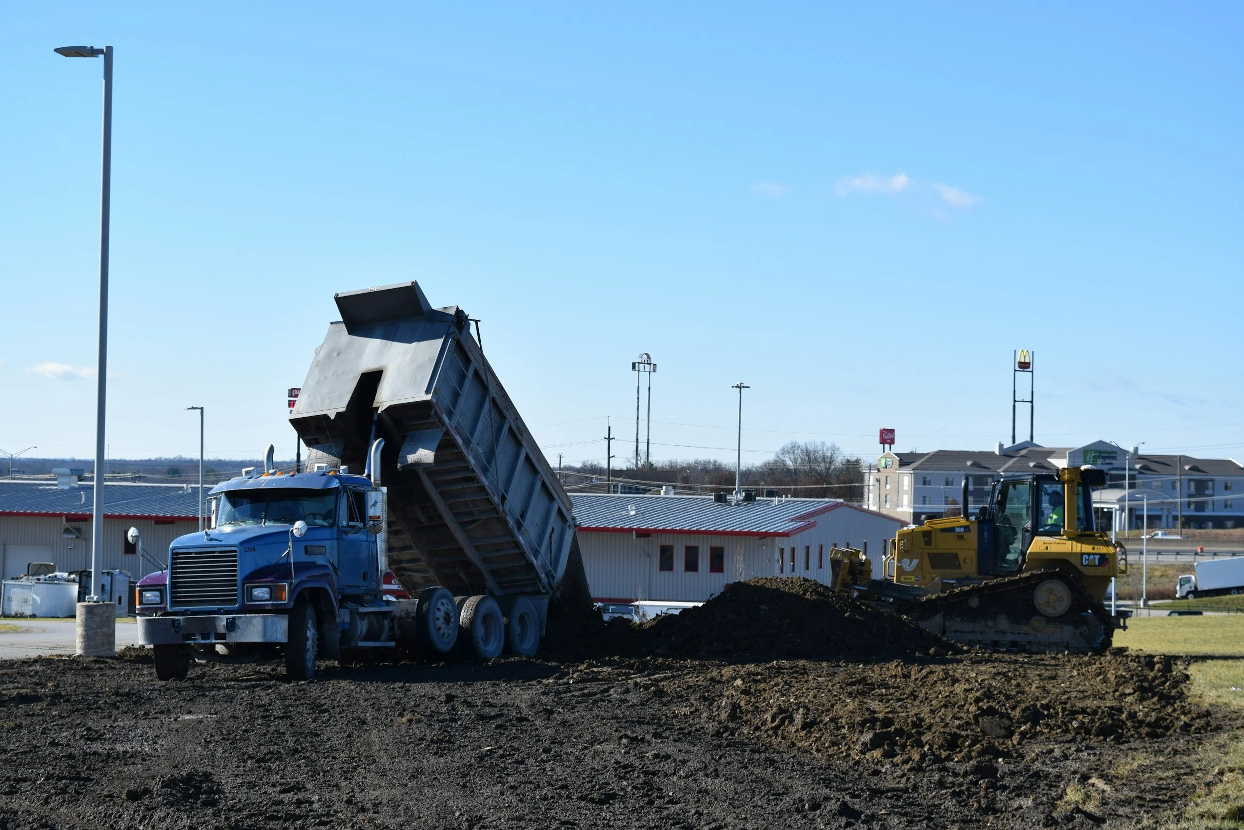 A construction site with a blue dump truck lifting its bed to unload soil, and a yellow bulldozer moving ground, under a clear blue sky.