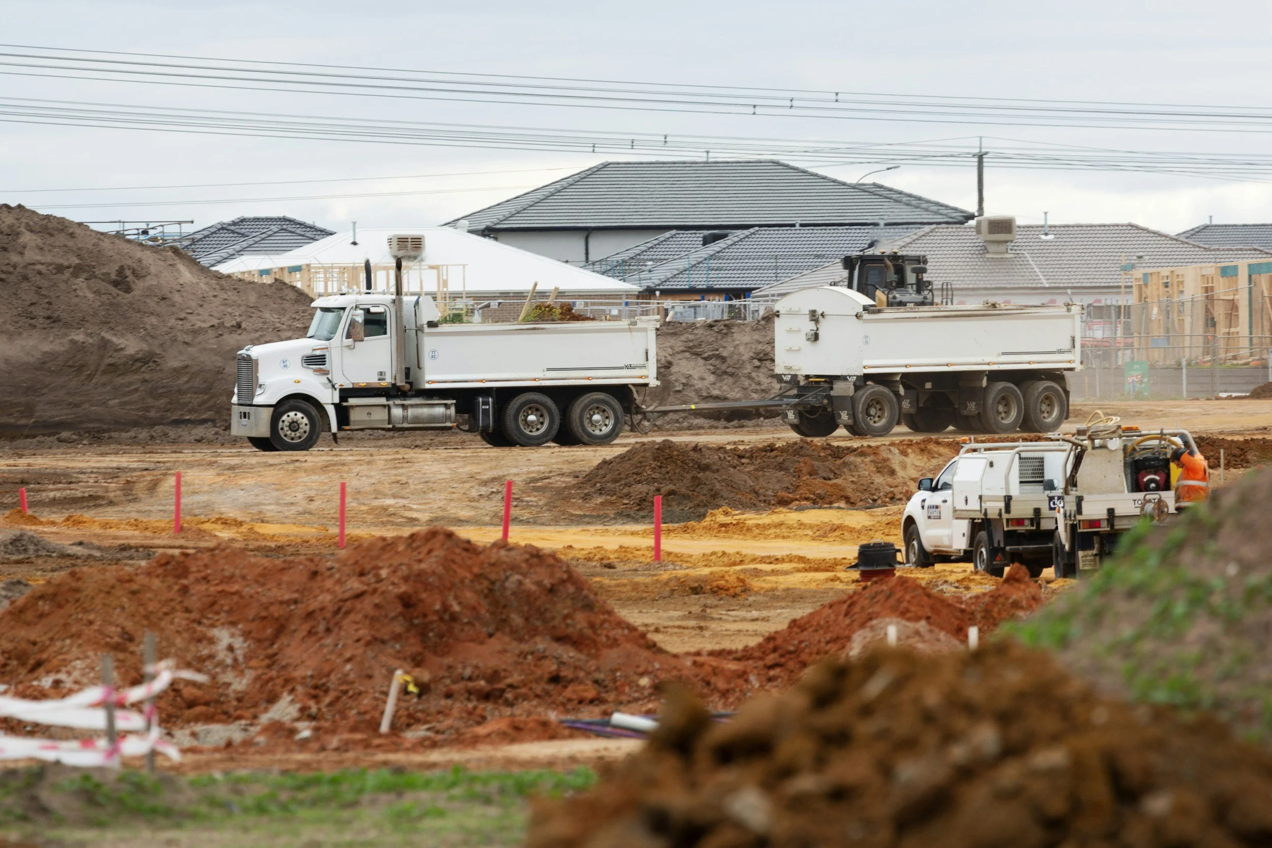 Construction site with trucks and workers, houses in the background, and construction materials and equipment in the foreground.
