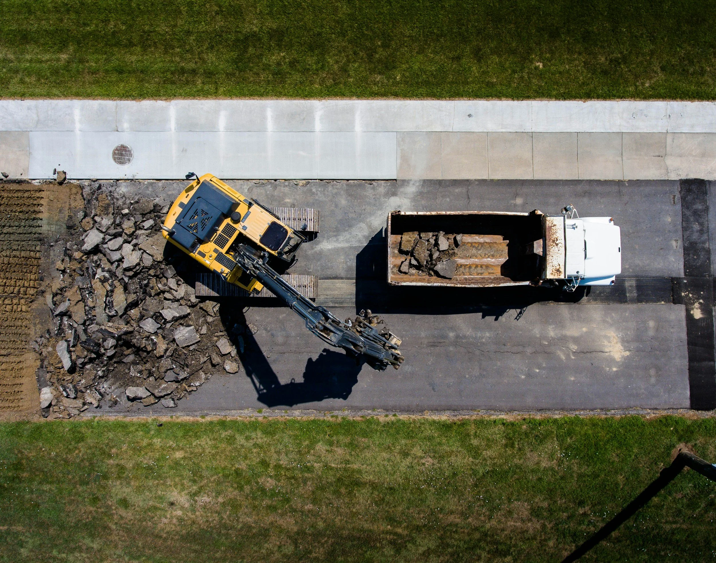 An aerial view of a construction site showing a cement sidewalk, a yellow construction vehicle, a dump truck with rocks, and green grass surrounding the area.
