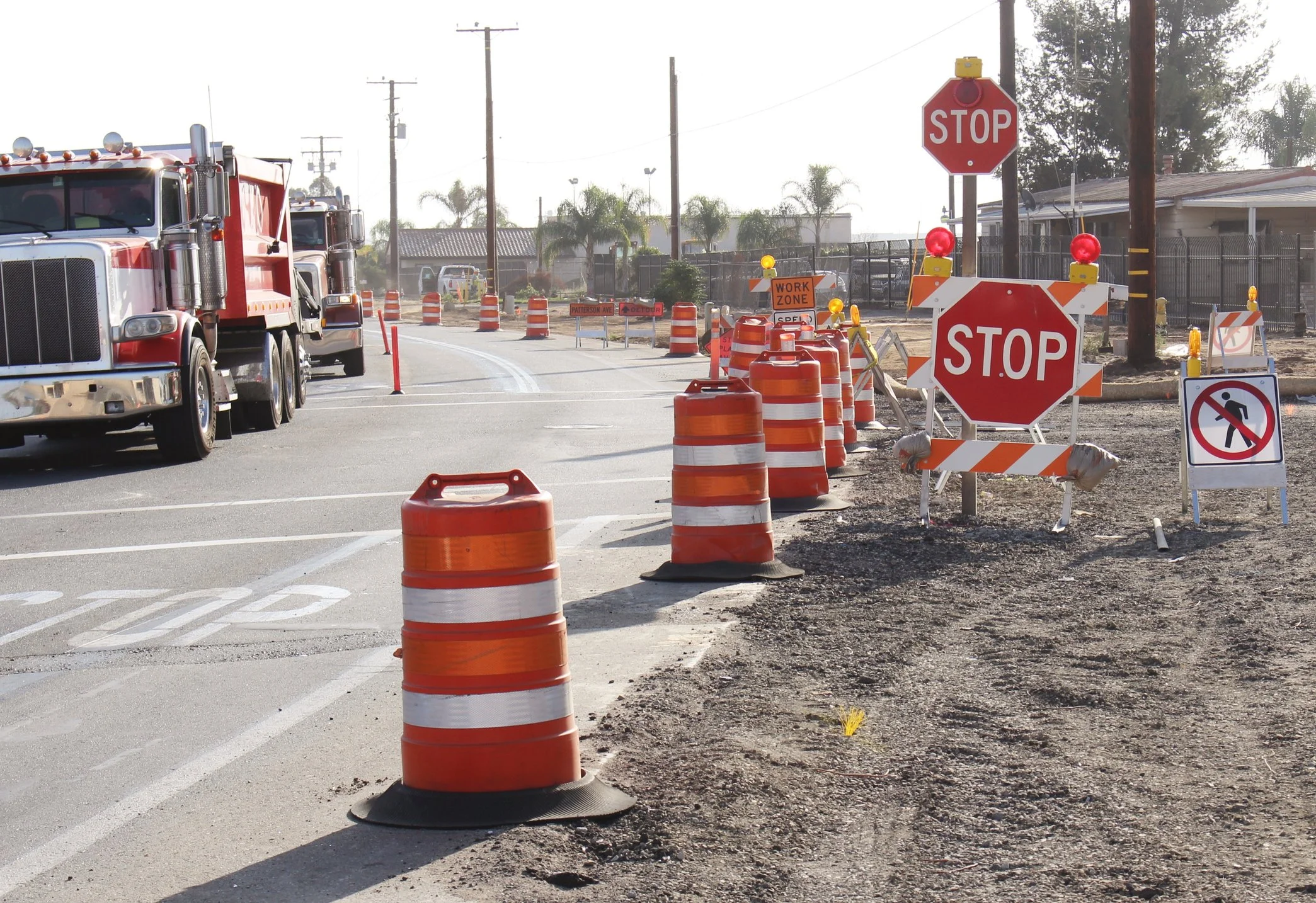 Road construction zone with orange barrels, stop signs, and warning signs, with trucks and construction equipment in the background.