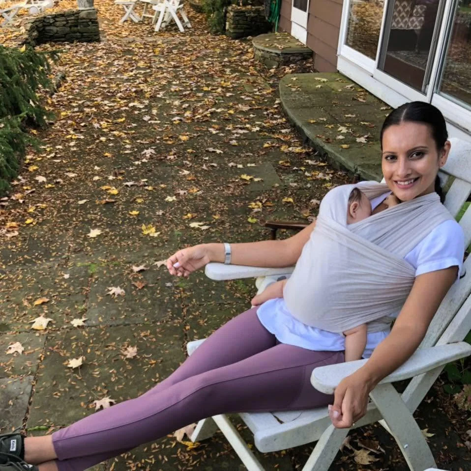 Sulekha sitting on a white outdoor chair with a smiling face, carrying a baby in a sling, in a backyard with fallen autumn leaves.