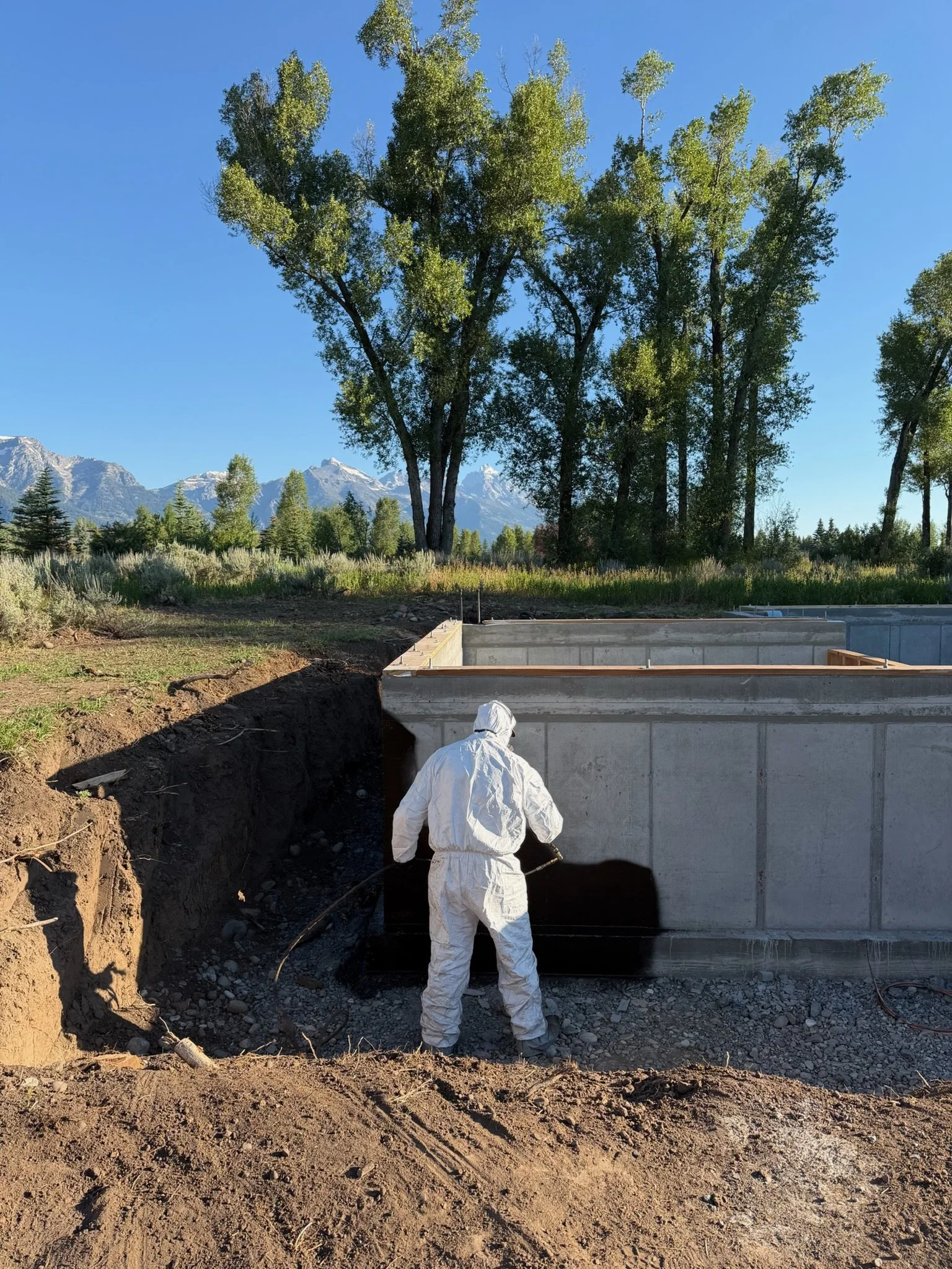 Person in white protective suit working at a construction site with foundation walls, outdoors, trees, mountains, and clear blue sky in the background.