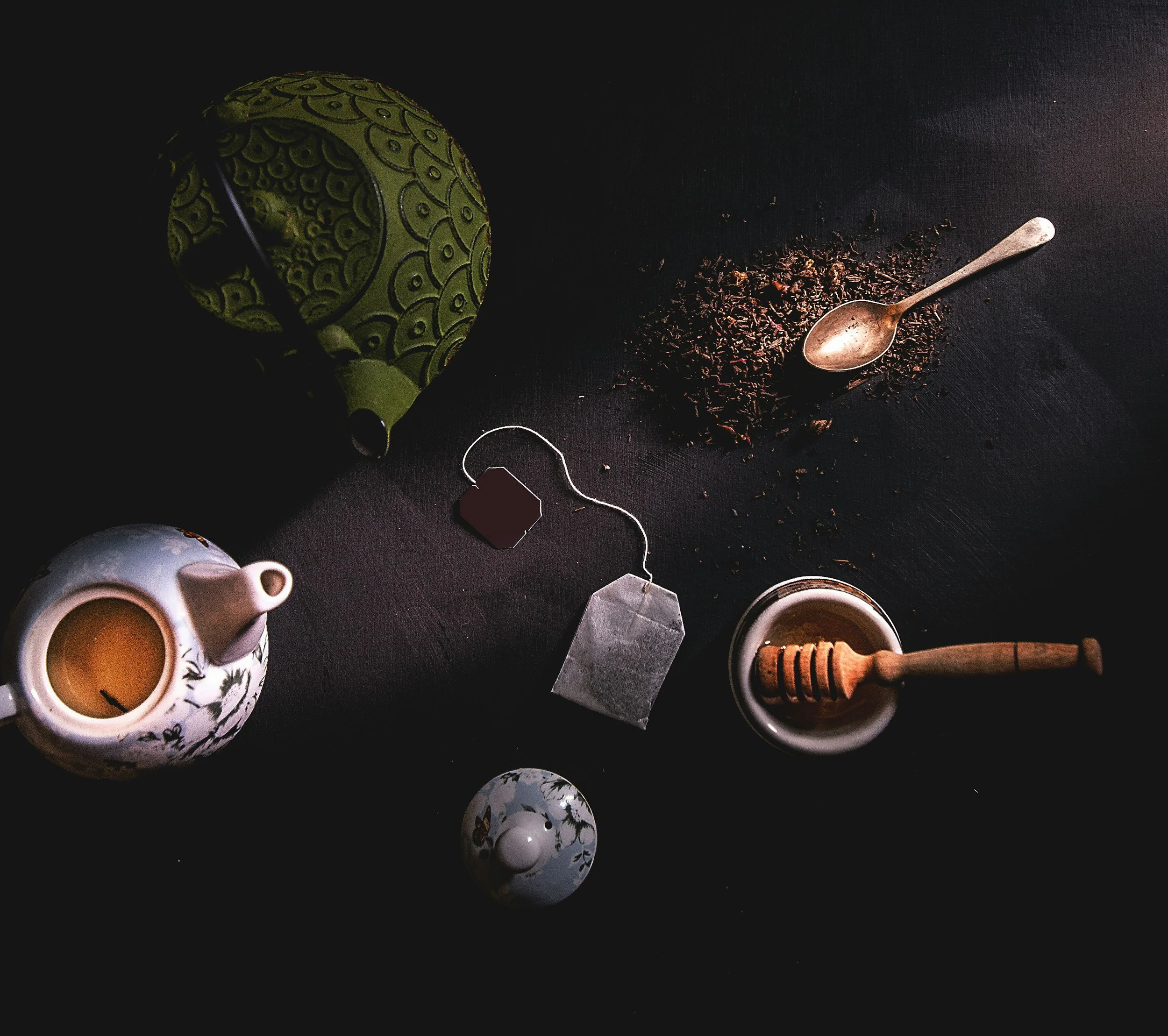 Close-up of a black surface with a green and black patterned hat, a spilled container of loose tea leaves, a silver spoon, a honey dipper, and two cups of tea.