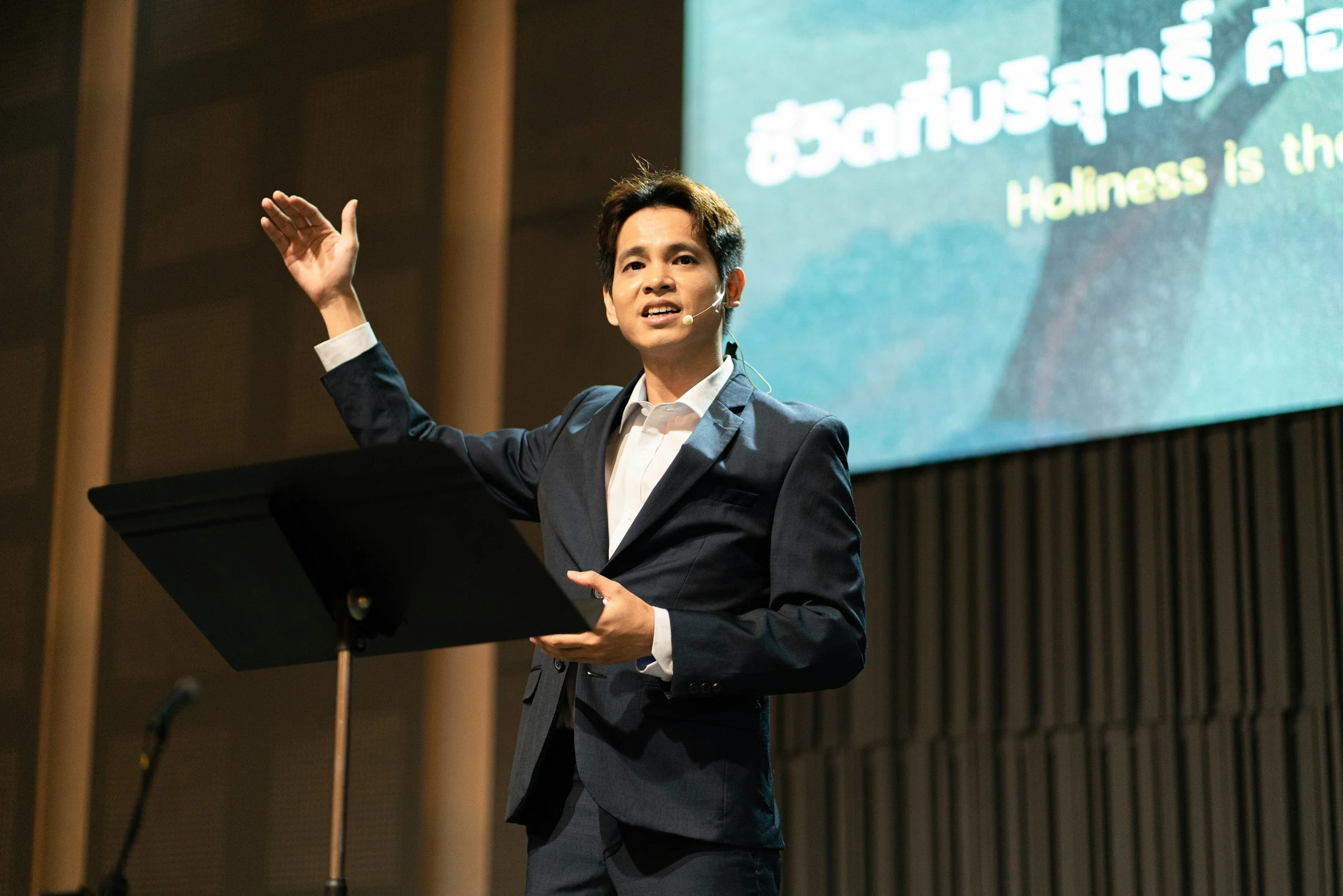 A man in a dark suit and white shirt giving a presentation with a microphone headset. He is standing at a podium with his right hand raised. A large screen behind him displays text in Thai and English, mentioning 'Holiness'.