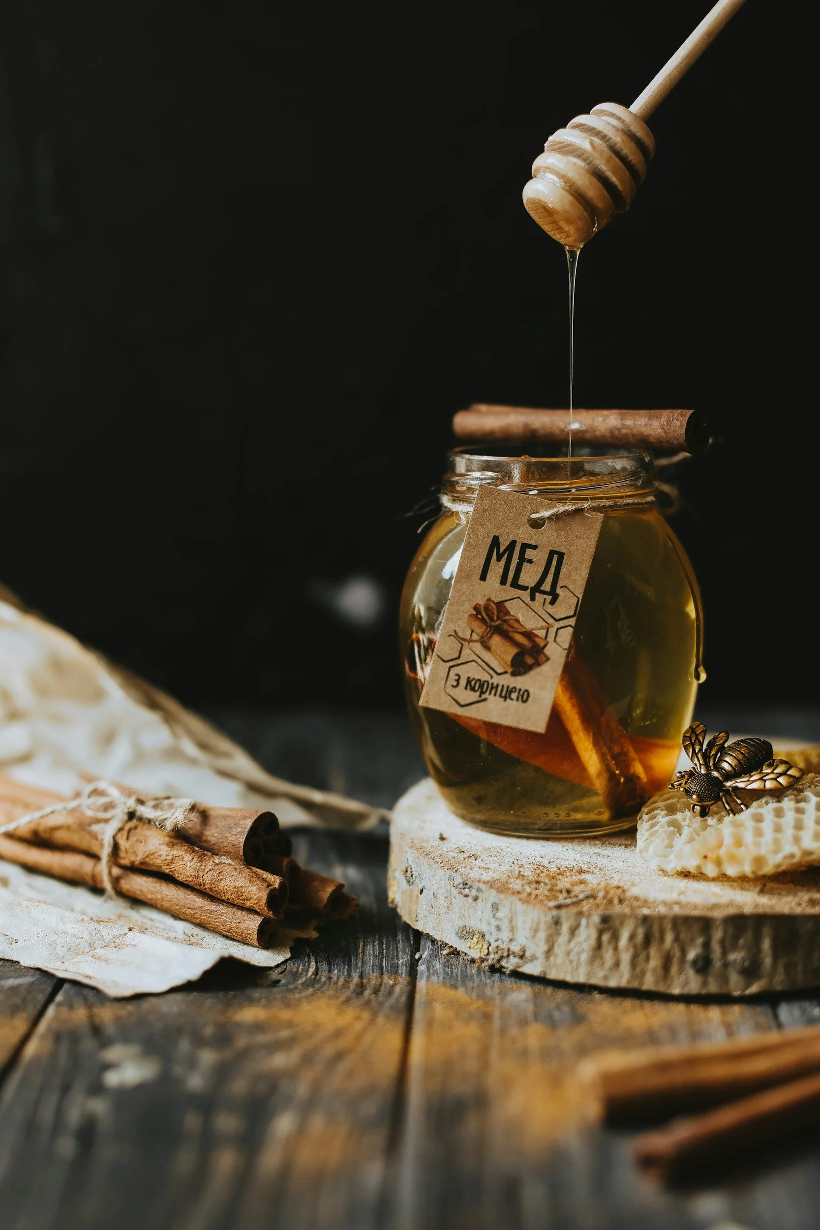 A jar of honey with a honey dipper above it, surrounded by cinnamon sticks, dried flowers, and honeycomb on a rustic wooden table.