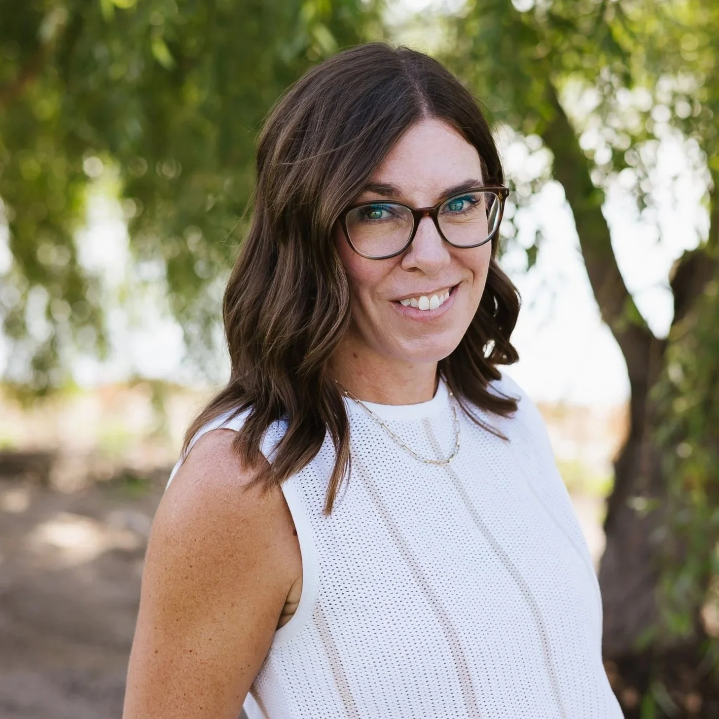A woman with shoulder-length brown hair, wearing glasses and a black-and-white striped shirt, smiling outdoors with green trees in the background.