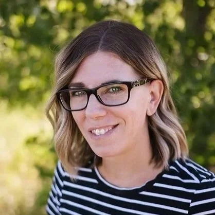 Woman with glasses and shoulder-length brown hair, wearing a black and white striped shirt, outdoors with greenery in the background.