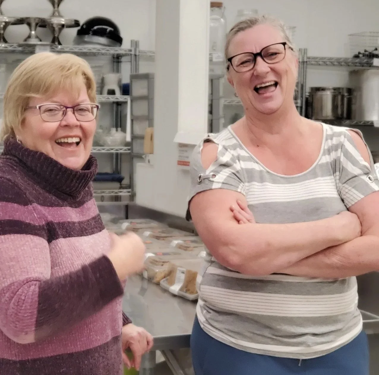 Two women smiling and laughing in a kitchen or food prep area, with shelves of kitchenware in the background.