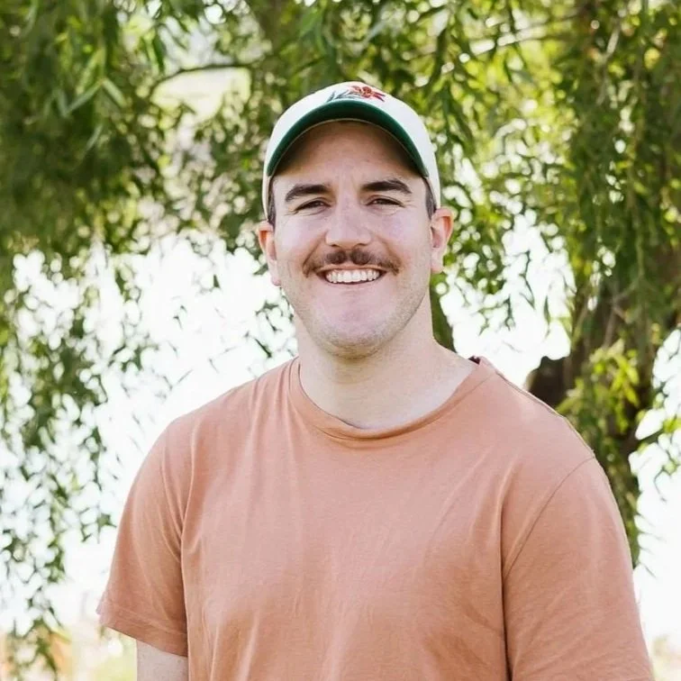 A smiling man wearing a black cap and an olive green shirt outdoors with blurred trees in the background.