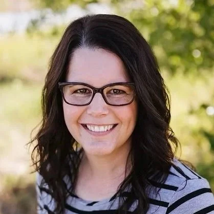 Woman with dark wavy hair and glasses smiling outdoors, wearing a striped shirt.