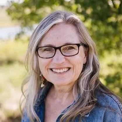 Smiling woman with glasses and long gray hair outdoors, wearing a denim jacket.