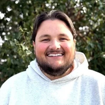 A smiling man with dark hair and a beard, wearing a light gray hoodie, standing outdoors in front of green leafy trees.