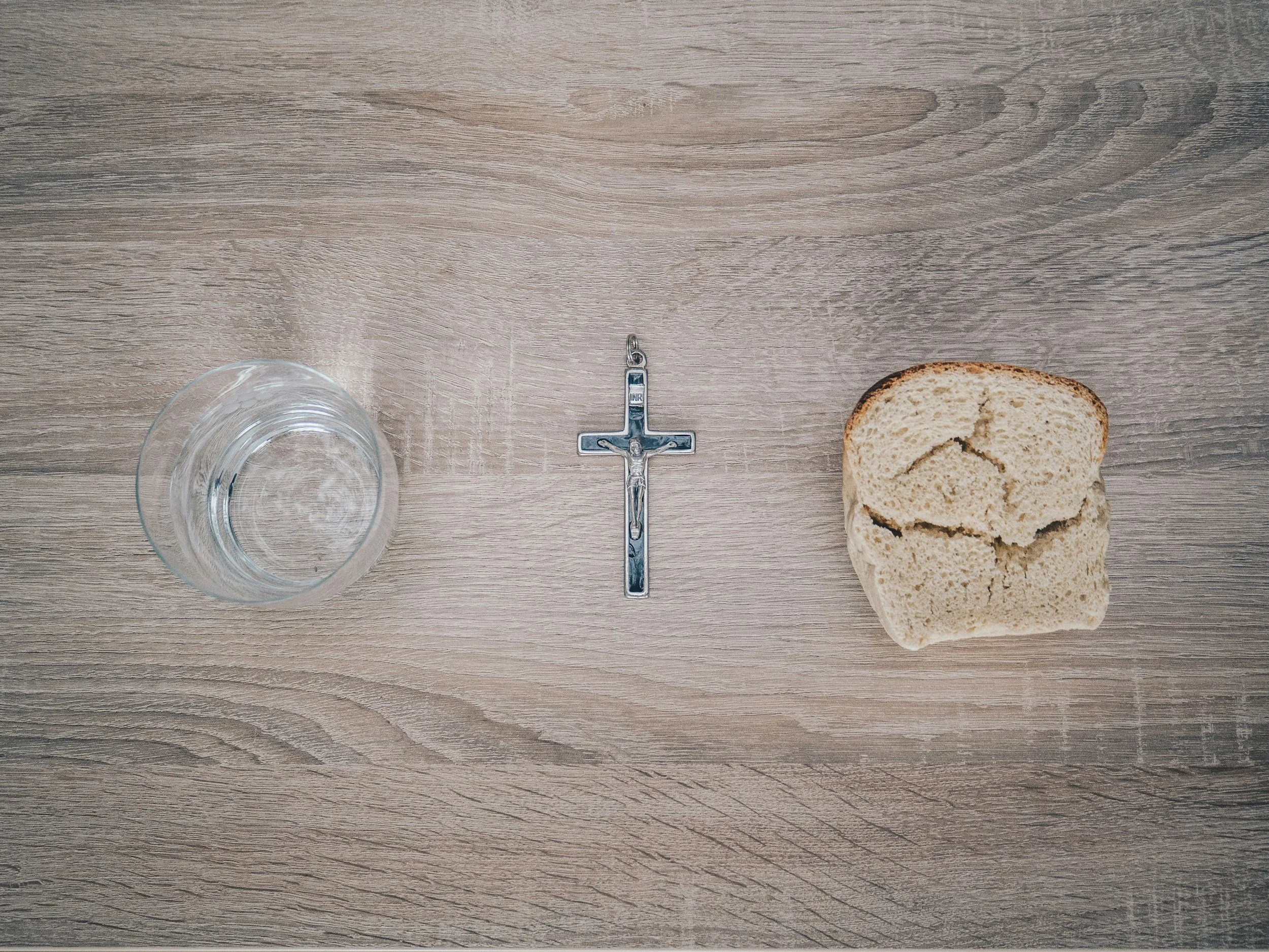 A transparent glass, a crucifix, a slice of bread, and a metal key on a wooden surface.
