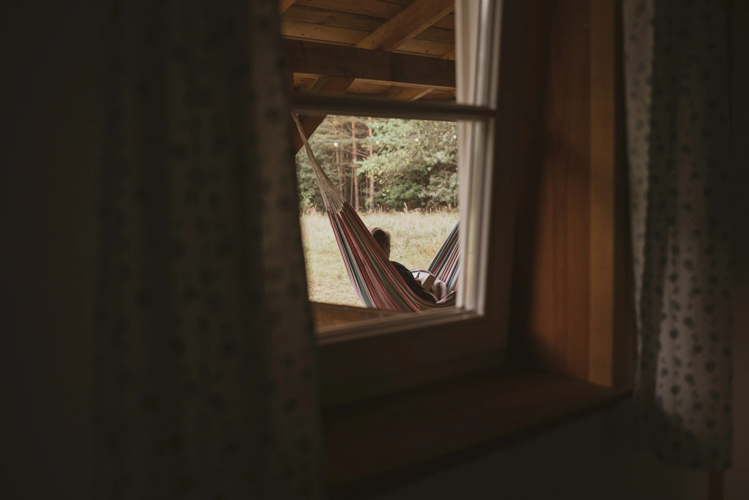 A person resting in a hammock outside a wooden cabin, viewed through a small window with curtains.