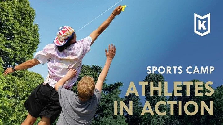 A girl wearing a hat and sports outfit is being lifted by a boy with blonde hair, reaching upward towards a discus during a sports camp outdoors under a blue sky with trees in the background.