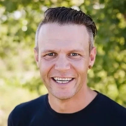 Smiling man with short dark hair in outdoor setting with green foliage in background.