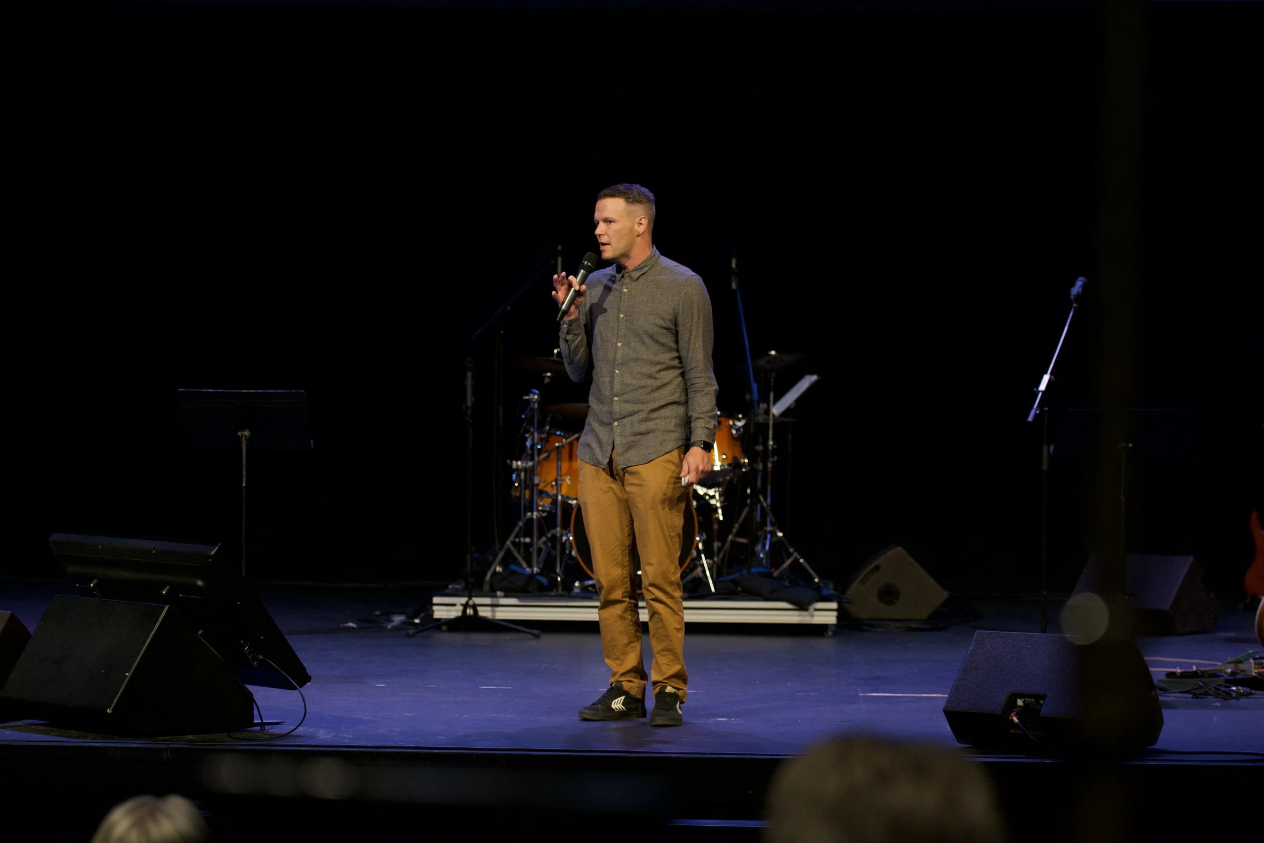 A man stands on stage holding a microphone, speaking or singing, with musical instruments including drums in the background, dim lighting.