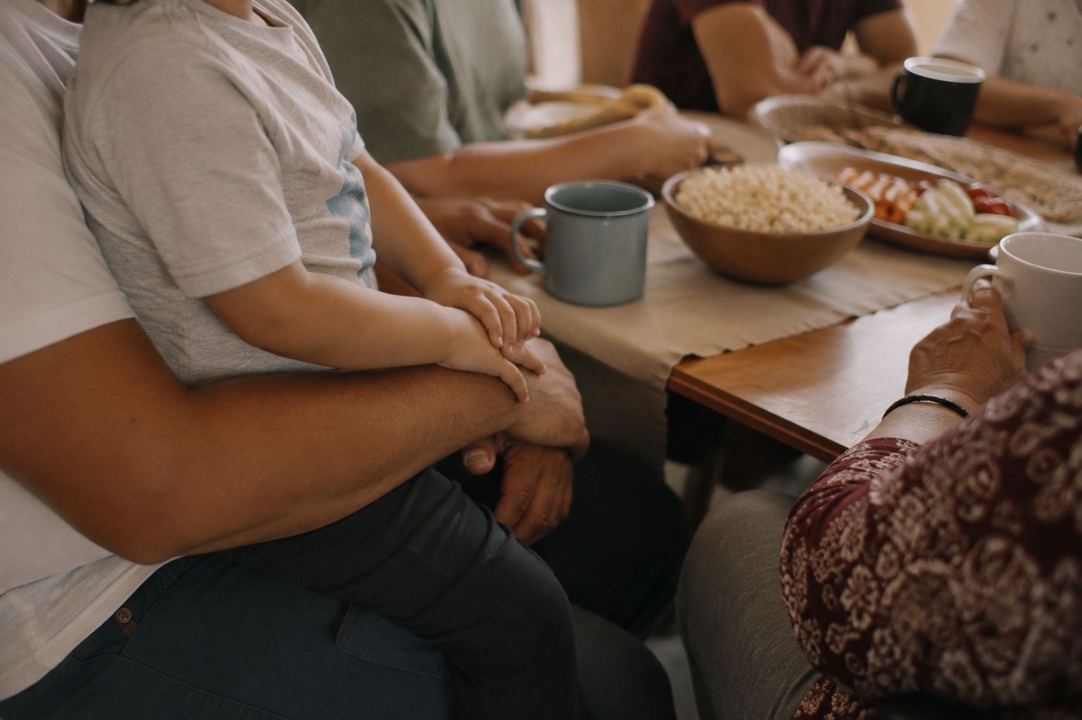 People sitting around a dining table with bowls of food and coffee mugs, including a child sitting on an adult's lap.