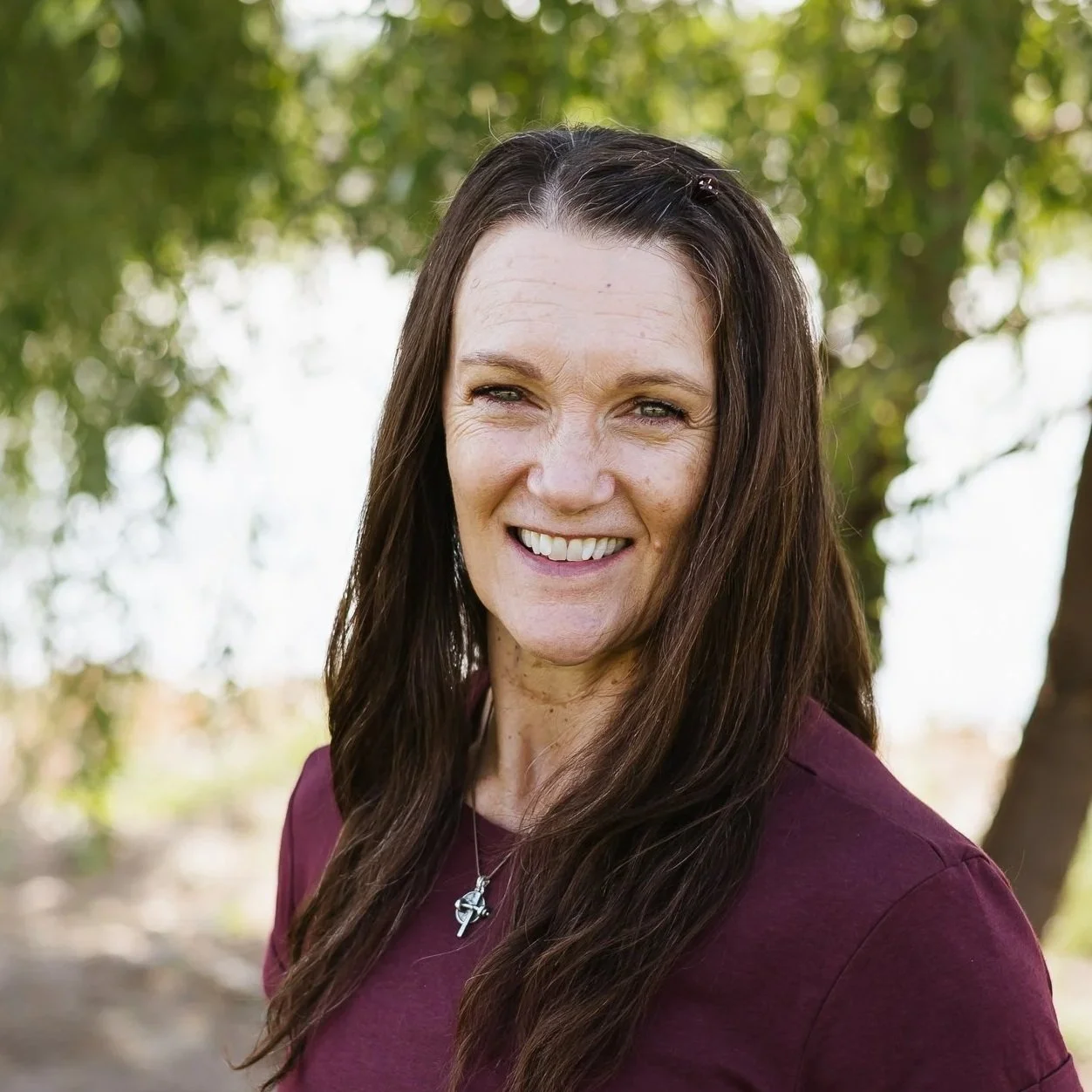 Smiling woman with long brown hair in a braid, outdoors with greenery background.