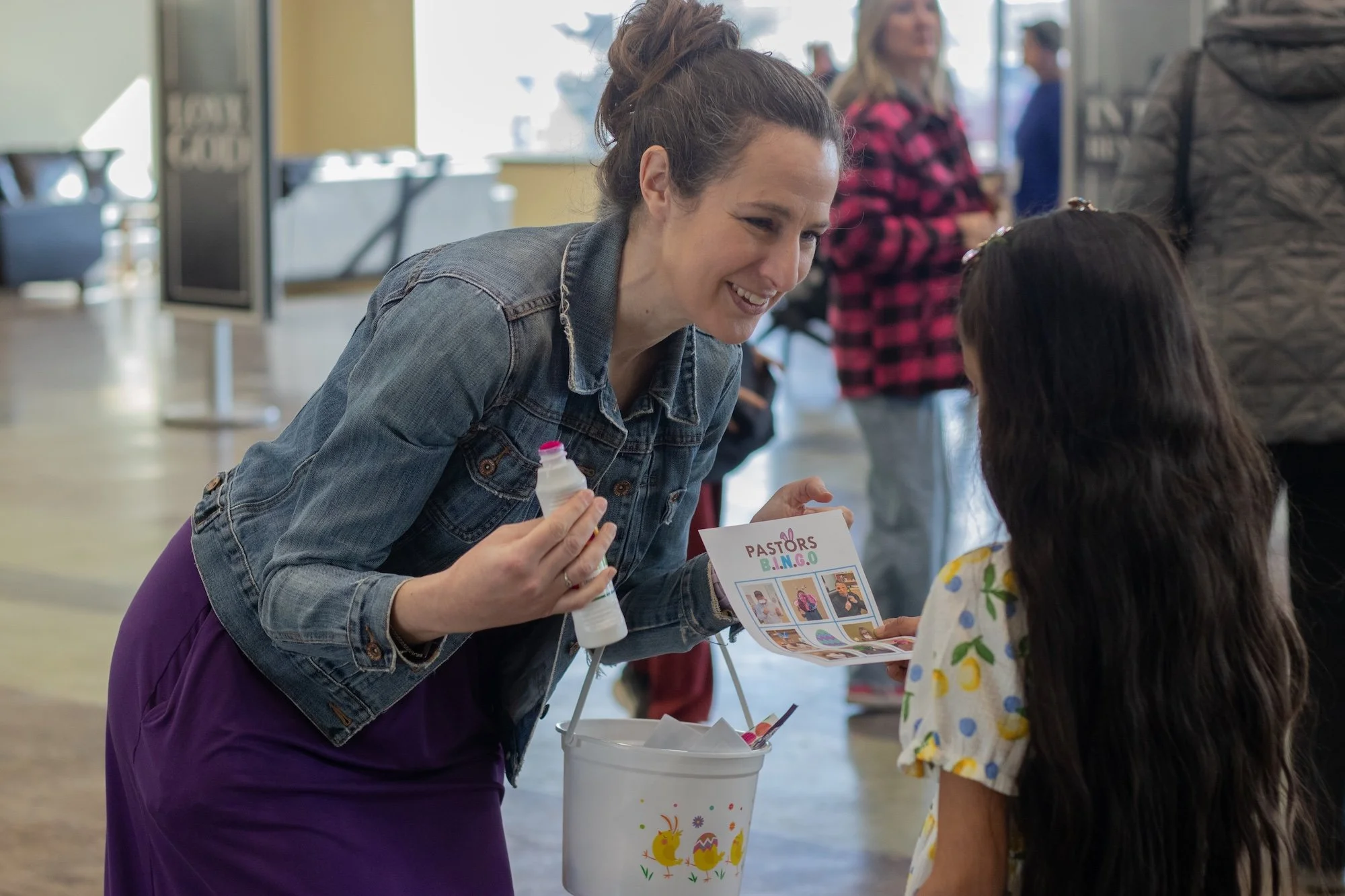 A woman with a bun hairstyle, wearing a denim jacket and purple dress, smiling as she interacts with a young girl at a community event. The woman holds a bottle of glue and a flyer with pictures, and there is a white bucket decorated with Easter eggs and chicks nearby. Other people are visible in the background inside a brightly lit indoor space.