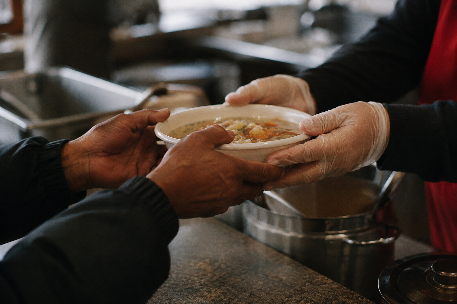 Person in black jacket receiving a bowl of soup from a worker wearing gloves in a kitchen or food pantry.