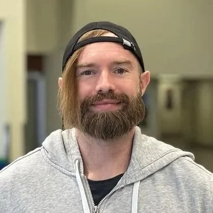 A man with a beard wearing a gray hoodie and a backward black baseball cap, smiling indoors.