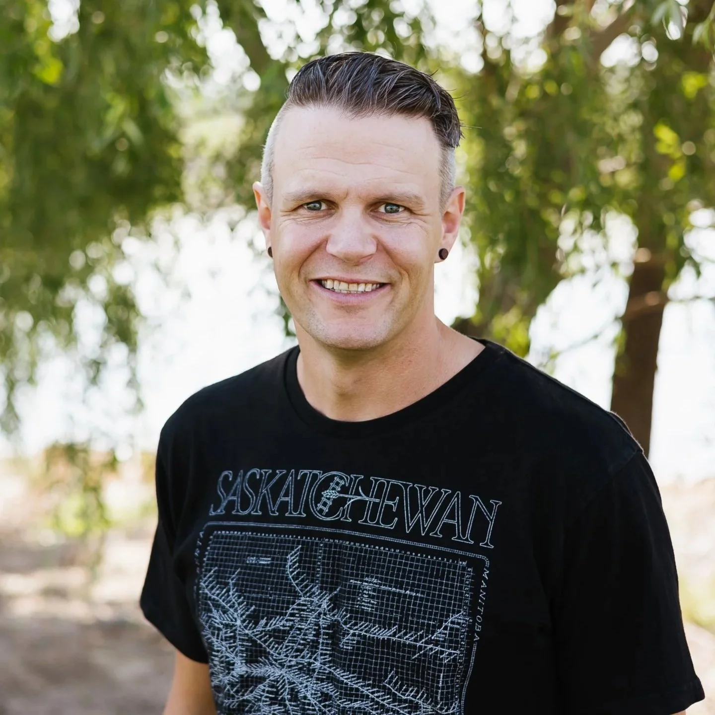 A smiling man with short, styled hair and earrings, outdoors with green trees in the background.