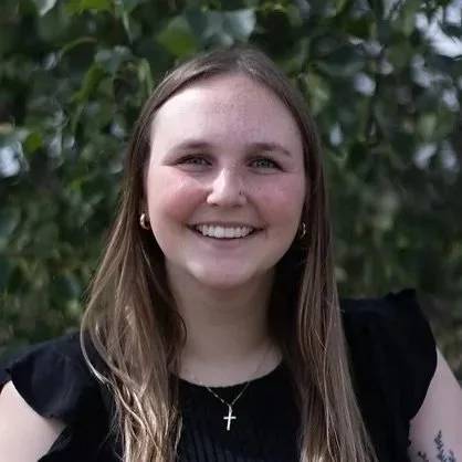 A smiling young woman with long brown hair, wearing a black top, earrings, a cross necklace, and standing outdoors with green foliage in the background.