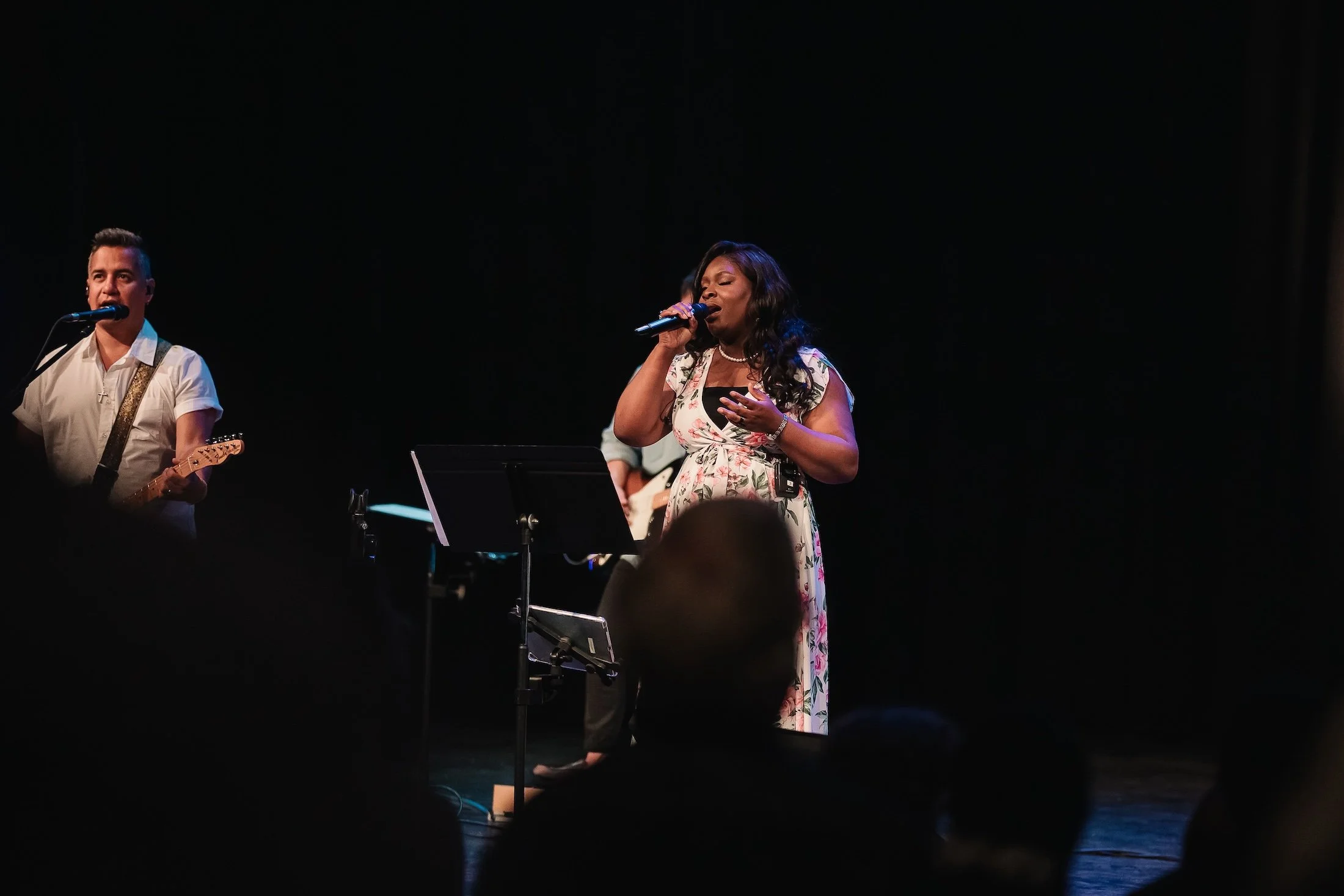 A female singer in a floral dress performing with a microphone on stage, accompanied by male guitar players in a dark setting.