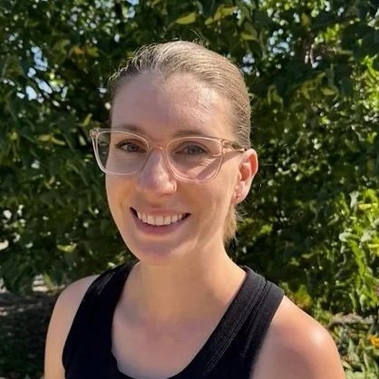 A woman with glasses and a black sleeveless top smiling outdoors with green foliage in the background.