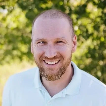 A smiling man with a beard and short hair, wearing a white collared shirt, outdoors with green foliage in the background.